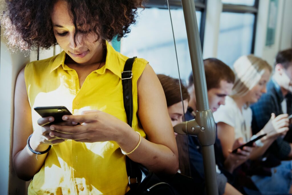 woman in yellow top standing on a train grabbed by the eyeballs on her mobile device