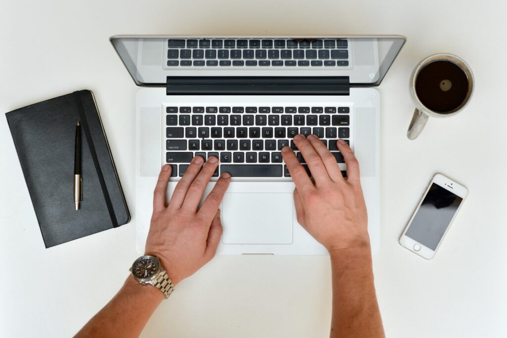 hands typing on a laptop on desk with notebook and coffee and phone