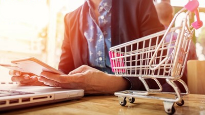 man reading how to start your own amazon business in 7 simple steps on his smartphone with a laptop and minature shopping cart on the desk beside him