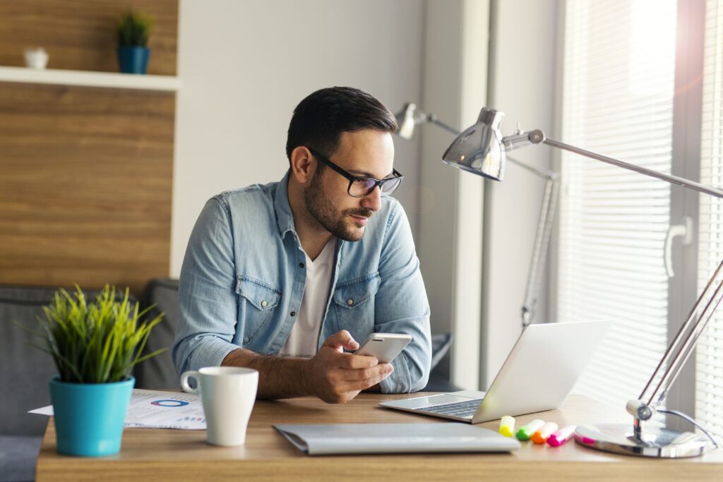 man wearing glasses and holding a smartphone sitting at desk reading how to start your own amazon business in 7 simple steps on a laptop