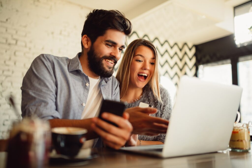 smiling couple sitting at table in cafe reading 5 ways to increase your business profits blog on their laptop