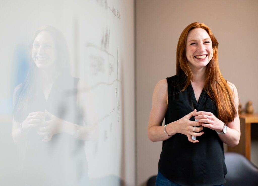 smiling red haired woman standing next to a whiteboard with a pen in her hand working on 3 simple strategies to boost her profits.
